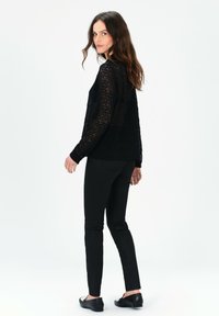 Top lungo a maniche con pizzo nero e motivo testurizzato, abbinato a pantaloni neri slim-fit e scarpe nere flat. Modella che guarda oltre la spalla.