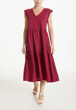 Donna che indossa un vestito midi bordeaux a più balze senza maniche, con scollo a V e tacchi bianchi con cinturino alla caviglia e punta aperta, in piedi su uno sfondo bianco.