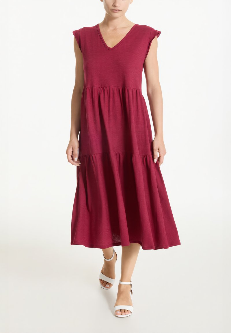 Donna che indossa un vestito midi bordeaux a più balze senza maniche, con scollo a V e tacchi bianchi con cinturino alla caviglia e punta aperta, in piedi su uno sfondo bianco.