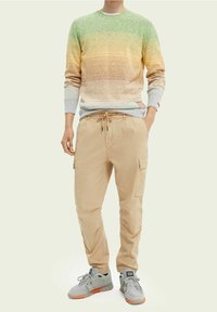 Person iført en sweater med gradient fra grøn til brun, beige cargo-bukser med snøre og grå sneakers med orange såler, står med afslappede hænder.