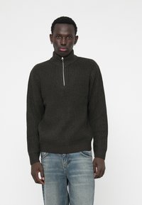 Dark green ribbed sweater with a high collar and half-zip closure, featuring textured knit and long sleeves, paired with light blue jeans.