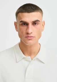 Valge polo särk, millel on volditud krae ja ees nööbid. Mudelil on lühikesed juuksed ja neutraalne ilme. Lihtne disain, ühtlane kangas.