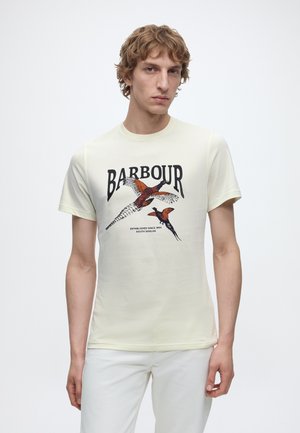 Jeune homme portant un t-shirt crème avec un graphisme de faisan en vol et le texte "Barbour", associé à un pantalon blanc, debout devant un fond uni.