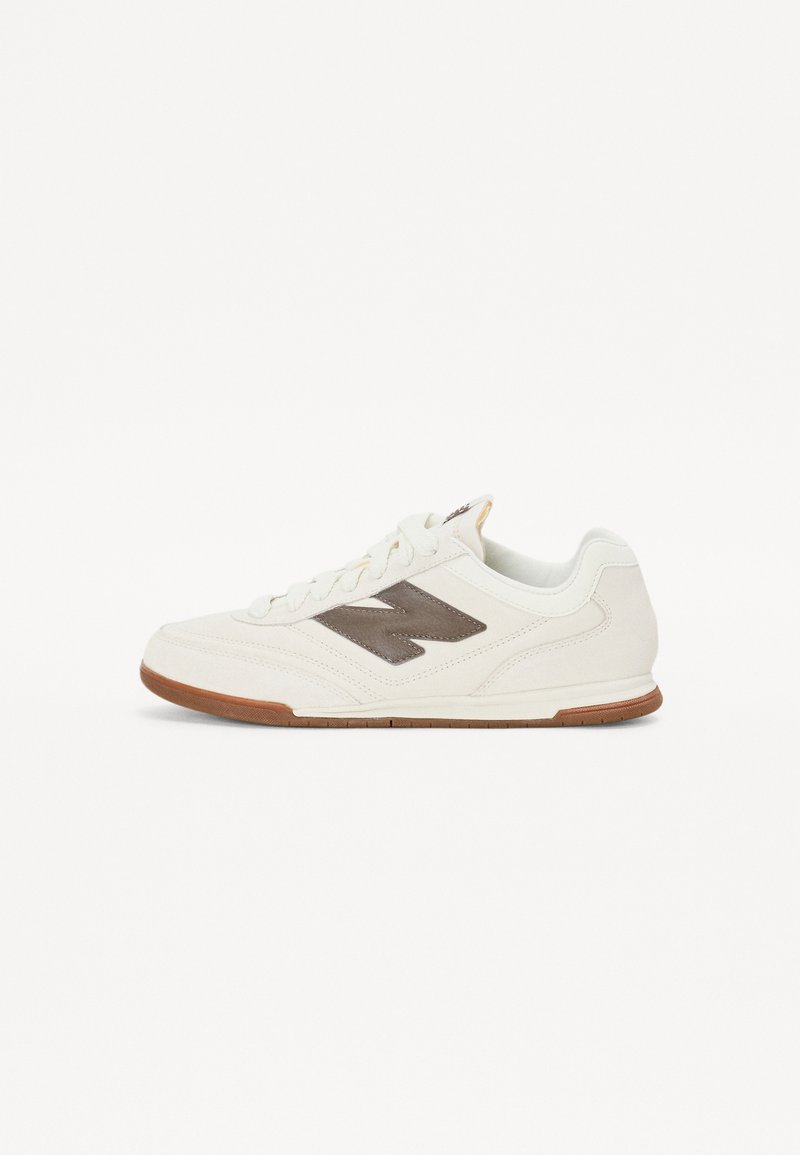 Baltas sneakers ar brūnu logotipu, izgatavoti no zamšādas un acs materiāla, ar noapaļotu purngalu, platu gumijas zoli un šņorēm. Vienkārša dizaina ar minimālām akcentēm.