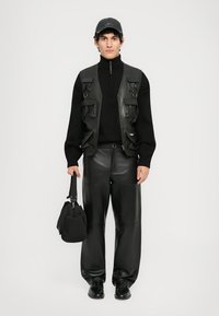 Black leather vest with multiple pockets, black turtleneck, and wide black leather pants. Holding a black bag, wearing a black cap.
