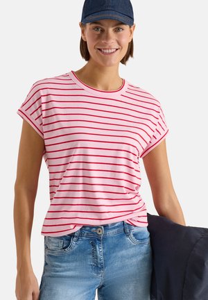 Uśmiechnięta kobieta w granatowej czapce, różowo-czerwonej bluzce w paski, jasno-niebieskich dżinsach, trzyma ciemną kurtkę na białym tle.