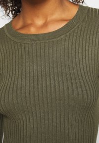 Gros plan sur une personne portant un pull en maille côtelée vert olive avec un col rond, dévoilant les clavicules et le haut de la poitrine.