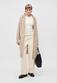 Long cardigan beige avec une capuche, tissu texturé, haut crème à manches longues, large pantalon clair et un sac tote noir. Chaussures noires.