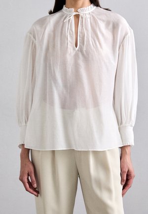 Personne portant un chemisier blanc transparent à manches longues avec un col montant à volants, une encolure à nouer, et un pantalon beige ample.
