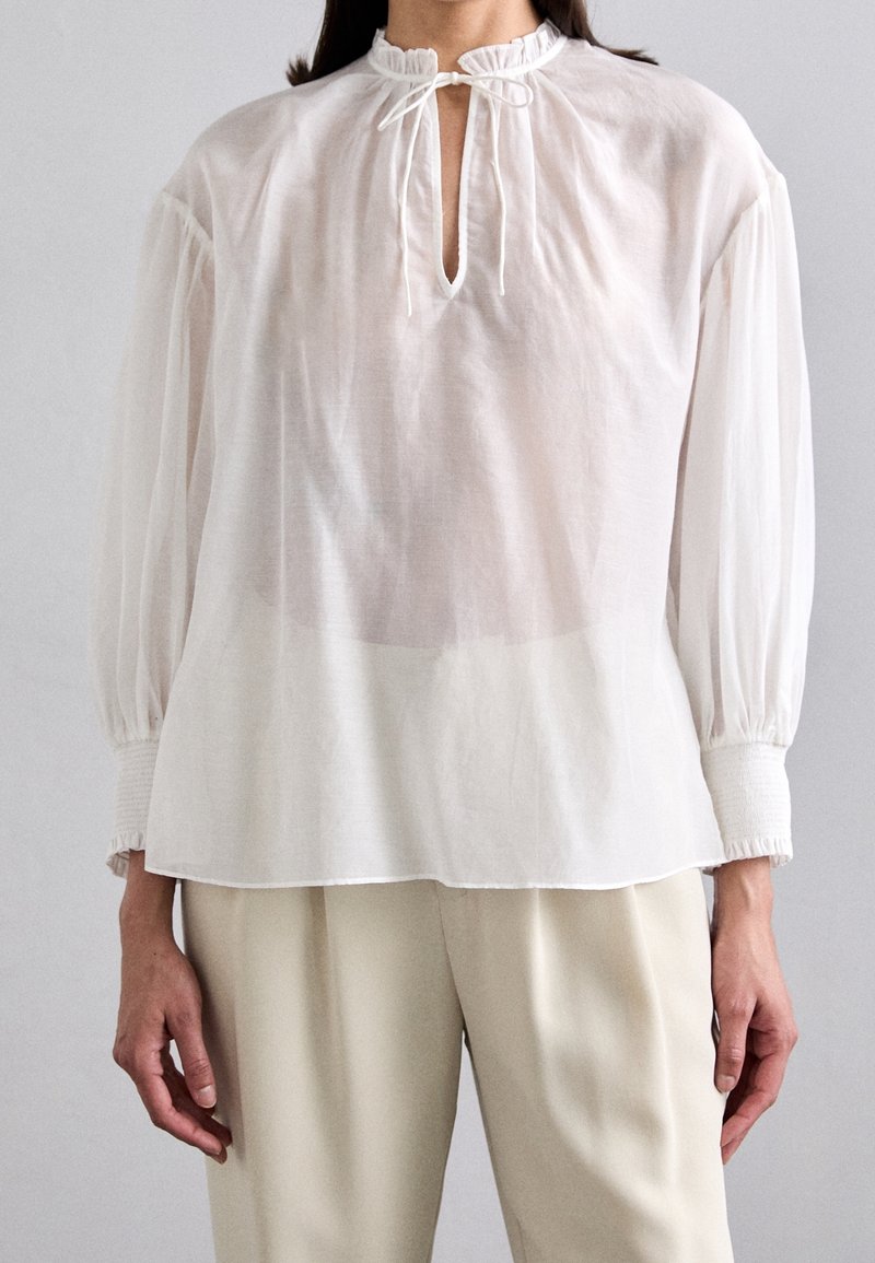 Personne portant un chemisier blanc transparent à manches longues avec un col montant à volants, une encolure à nouer, et un pantalon beige ample.