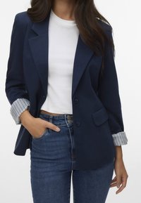 Marinblå blazer med en strukturerad design, två knappar och en framficka, med randiga manschettar. Kombinerad med en vit topp och blå jeans.