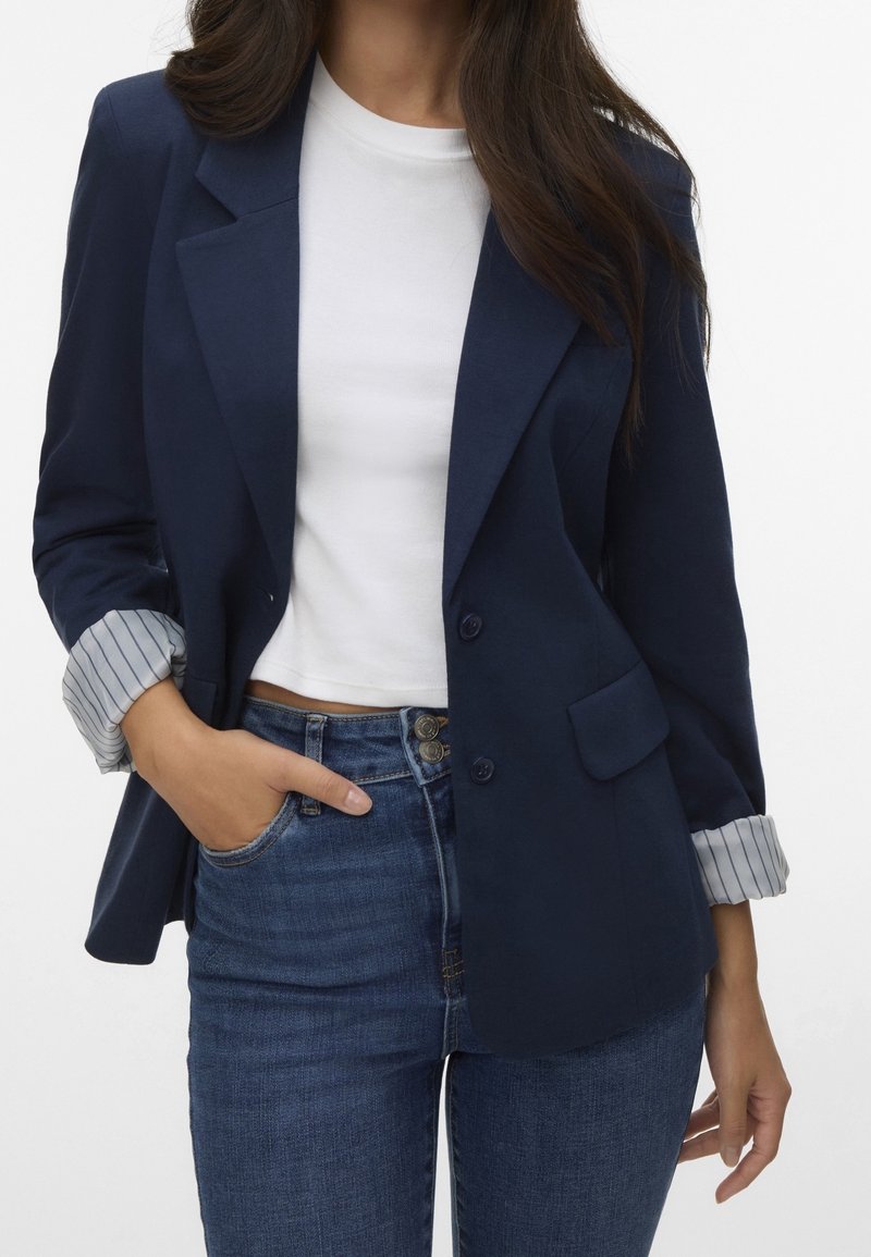 Marinblå blazer med en strukturerad design, två knappar och en framficka, med randiga manschettar. Kombinerad med en vit topp och blå jeans.