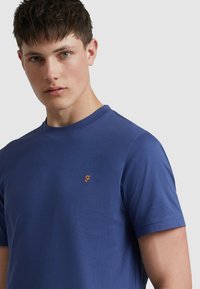 T-shirt di cotone blu con scollo rotondo, maniche corte e un piccolo logo ricamato arancione sul lato sinistro del petto. Texture liscia.