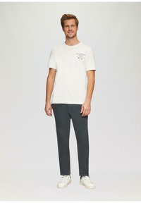 Witte katoen t-shirt met blauwe grafische tekst op de linkerkant van de borst, gecombineerd met donkergrijze broek en witte sneakers. Eenvoudige, casual outfit.