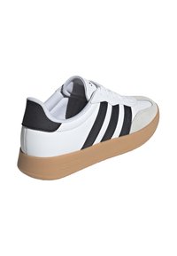 Chaussure de sport blanche avec des rayures noires, un bout en suède et une semelle en caoutchouc. Elle possède un col rembourré et une fermeture à lacets.