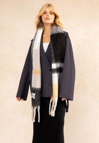 Écharpe multicolore au toucher doux et duveteux, avec des blocs noirs, gris, blancs et crème, et des bords à franges. Portée de manière lâche.