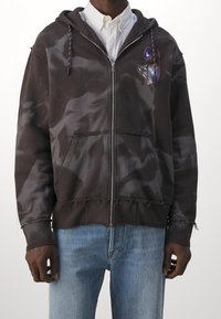 Sudadera con capucha y cremallera en marrón oscuro con un patrón de tie-dye, dobladillo desgastado y bolsillo frontal; presenta acentos gráficos coloridos en el pecho.