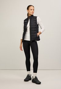 Schwarze Pufferweste mit hohem Kragen, Reißverschluss vorne und seitlichen Taschen. getragen über einem weißen Langarmoberteil und kombiniert mit schwarzen Leggings.