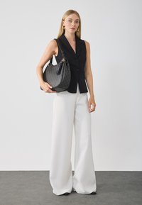Blazer sans manches noir avec des boutons, associé à un pantalon large blanc. Le modèle tient un sac à main texturé gris avec un accent en frange.