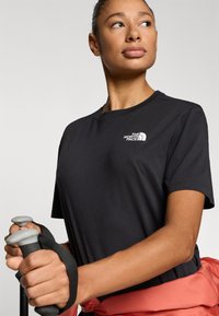 Μαύρο κοντομάνικο μπλουζάκι με λευκό λογότυπο, συνδυασμένο με γκρι μπαστούνια πεζοπορίας. Η μπλούζα έχει στρογγυλή λαιμόκοψη και άνετη εφαρμογή.