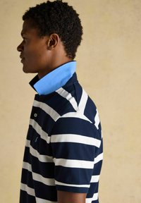 Homme portant un polo rayé bleu marine et blanc avec un col bleu clair relevé, face à gauche sur un fond beige uni.