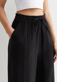 Zwarte, gestructureerde, wijde broek met een elastische tailleband en een trekkoord. Beschikt over zijzakken en een gekreukt stoffen ontwerp.