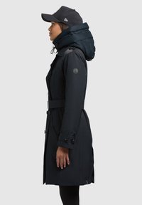 Cappotto lungo blu navy con cappuccio voluminoso, dotato di una vita con cintura, polsini con bottoni e una toppa con logo sulla manica. Abbinato a un berretto nero.