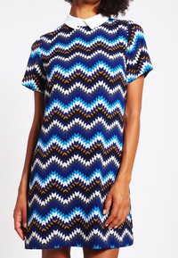Vestido de manga corta con un collar blanco. Presenta un patrón en zigzag en azul, marino y marrón. Hecho de una tela suave, diseño hasta la rodilla.