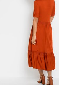 Rudy pomarańczowa midi sukienka z krótkimi rękawami, okrągłym dekoltem i warstwowym, falbaniastym dołem; wykonana z miękkiego, lekkiego materiału; charakteryzuje się luźnym krojem.