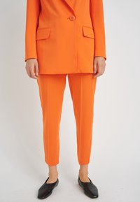 Oranje pak met een getailleerde blazer met één knoop en twee voorzakken, gecombineerd met smalle broeken. Zwarte instappers maken de look compleet.