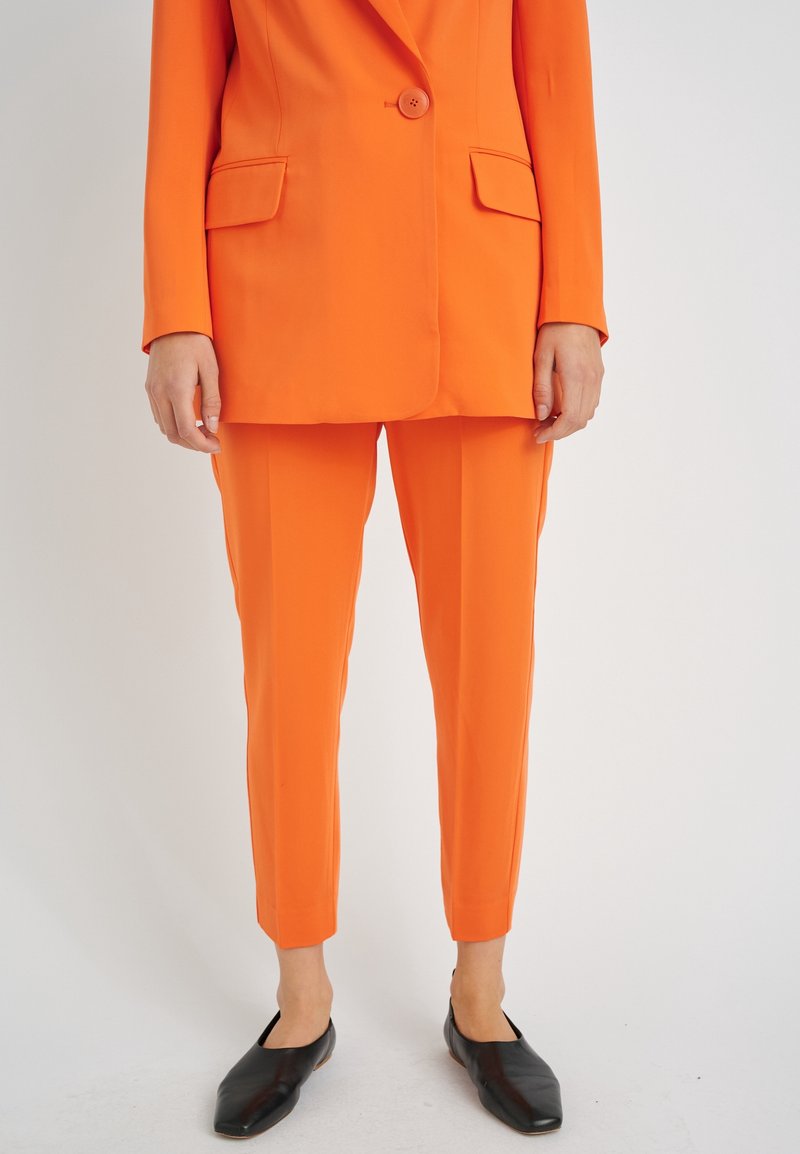 Oranje pak met een getailleerde blazer met één knoop en twee voorzakken, gecombineerd met smalle broeken. Zwarte instappers maken de look compleet.