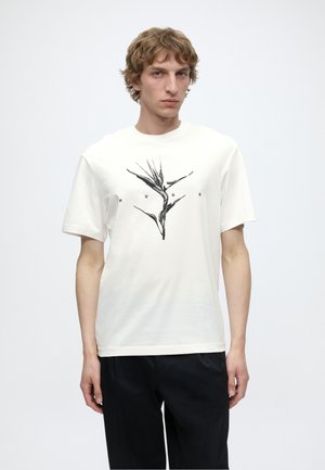 Jeune homme portant un T-shirt blanc avec un motif de fleur d'oiseau de paradis noir et les lettres "HUGO", associé à un pantalon noir, debout sur fond blanc.