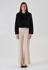 Veste courte en fausse fourrure noire à manches longues, associée à un pantalon beige taille haute à jambes larges, agrémenté de boutons dorés.