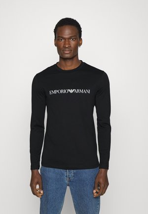 Tricou negru cu mânecă lungă, guler rotund și logo-ul alb "EMPORIO ARMANI" pe piept, purtat cu blugi albaștri.