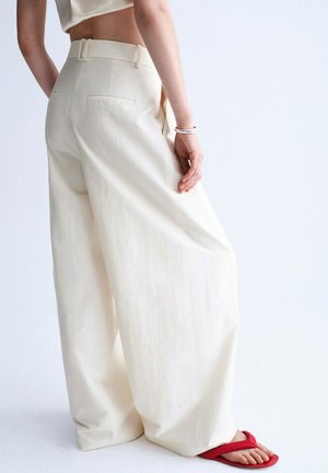 Person trägt hochtaillierte, weitgeschnittene cremefarbene Hosen, kombiniert mit roten Flip-Flops, und steht vor einem schlichten weißen Hintergrund.