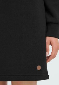 Zwart sweatshirt met een gladde textuur, voorzien van een ribgebreide manchette. Een klein, rond, bruin leren logo accent is zichtbaar nabij de zoom.