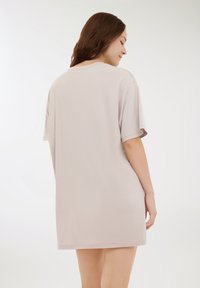 Donna con i capelli lunghi e castani, che indossa un vestito corto a maniche corte di un leggero color beige, in piedi di lato con la schiena parzialmente rivolta verso la telecamera.