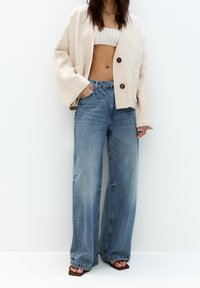 Vrouw in wijdvallende vervaagde blauwe jeans, een beige oversized jas met grote knopen, een gestreept cropped top en open teen bruine sandalen.