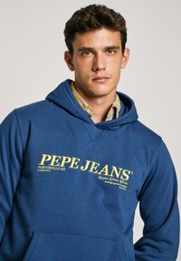 Modra kapuca iz mešanice bombaža z sprednjim žepom, nastavljivo kapuco z vrvico in rumenim logotipom na prsih.