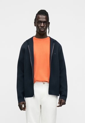 Sweat à capuche zippé bleu marine sur un t-shirt orange à col rond, associé à un pantalon blanc. Présente une texture lisse et un logo discret sur le sweat à capuche.