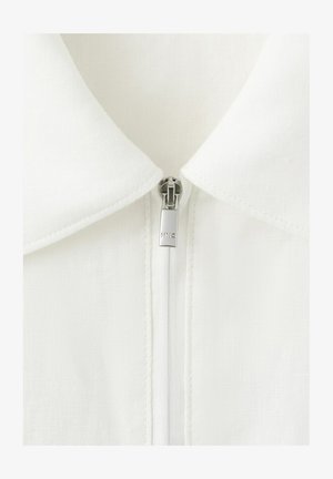 Witte stoffen kraag met een zilveren ritslipje gegraveerd met "MNG," gecentreerd en helemaal dichtgeritst tot aan de bovenkant van de kraag.