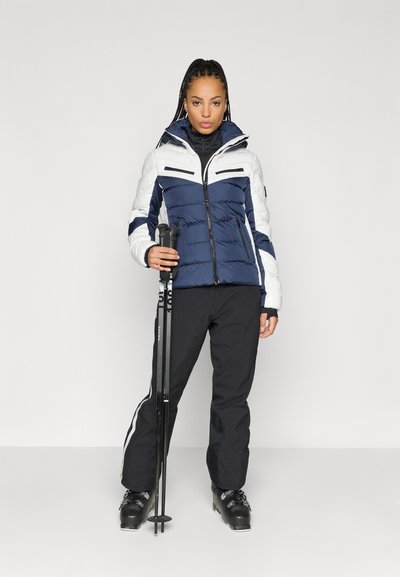 Μπλε και άσπρο αφράτο μπουφάν με οριζόντια ραφτά σχέδια, μαύρες παντελόνες σκι και μπαστούνια σκι. Λείο ύφασμα με φερμουάρ και λεπτομέρειες.