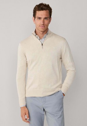 Hombre con suéter beige de media cremallera sobre camisa a rayas, pantalones gris claro, de pie frente a un fondo liso gris claro.