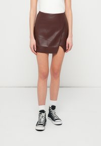 Μίνι φούστα από ψεύτικο δέρμα σε μπορντό απόχρωση, με διαγώνια περιτύλιξη, λεία υφή και στρίφωμα πάνω από το μέσο του μηρού, σε συνδυασμό με μαύρα αθλητικά παπούτσια.