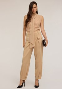 Blusa beige senza maniche con dettaglio a fiocco e perline decorative, abbinata a pantaloni beige high-waisted con tasche laterali. Scarpe nere con tacco.