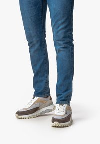 Sneakers sportive in grigio, beige e bianco con pannelli testurizzati e una suola spessa; indossati con jeans blu. Lacci e dettagli visibili.