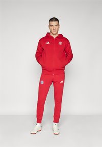 Tuta rosso composto da una felpa con zip e pantaloni elasticizzati. Logo Adidas e stemma del Bayern Monaco sul petto e sulla coscia. Scarpe da ginnastica bianche.
