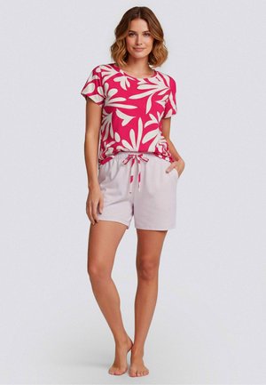 Kvinde står barfodet iført en pink og hvid blomsterprint kortærmet top og lyserøde shorts med snørre foran mod en ensfarvet baggrund.