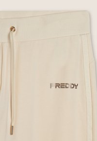 Joggers de color beige claro con una cinturilla acanalada, cordón y un logo plateado. El detalle lateral presenta una franja de marca en un tono similar. Material de algodón suave.