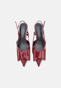 Ζευγάρι γυαλιστερών κόκκινων γόβων slingback με尖τρυπιά διοσκοποδων πίσω και μεγάλες διακοσμητικές κορδέλες σε λευκό φόντο.